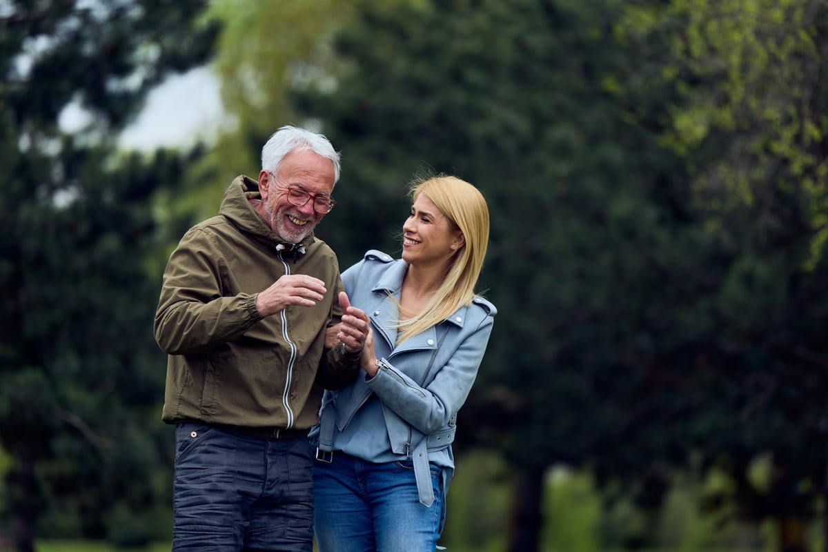 An older father and his adult daughter walking together through a sunny green park