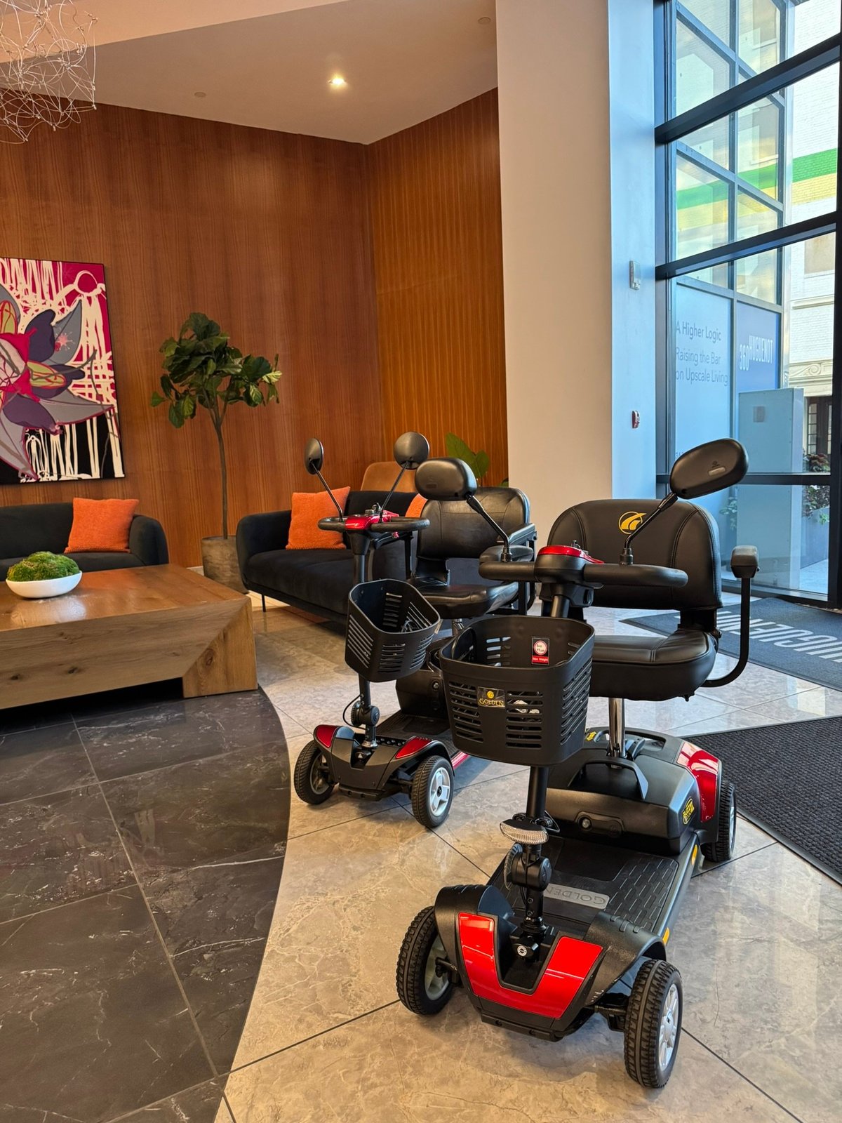 A red and black four-wheel Golden Buzzaround XL mobility scooter in the foreground of a modern lobby with wood-paneled walls and large windows