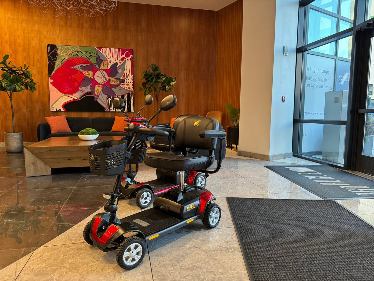 A red and black four-wheel Pride Go-Go Elite Traveller Plus mobility scooter parked in a hotel lobby with wooden paneled walls